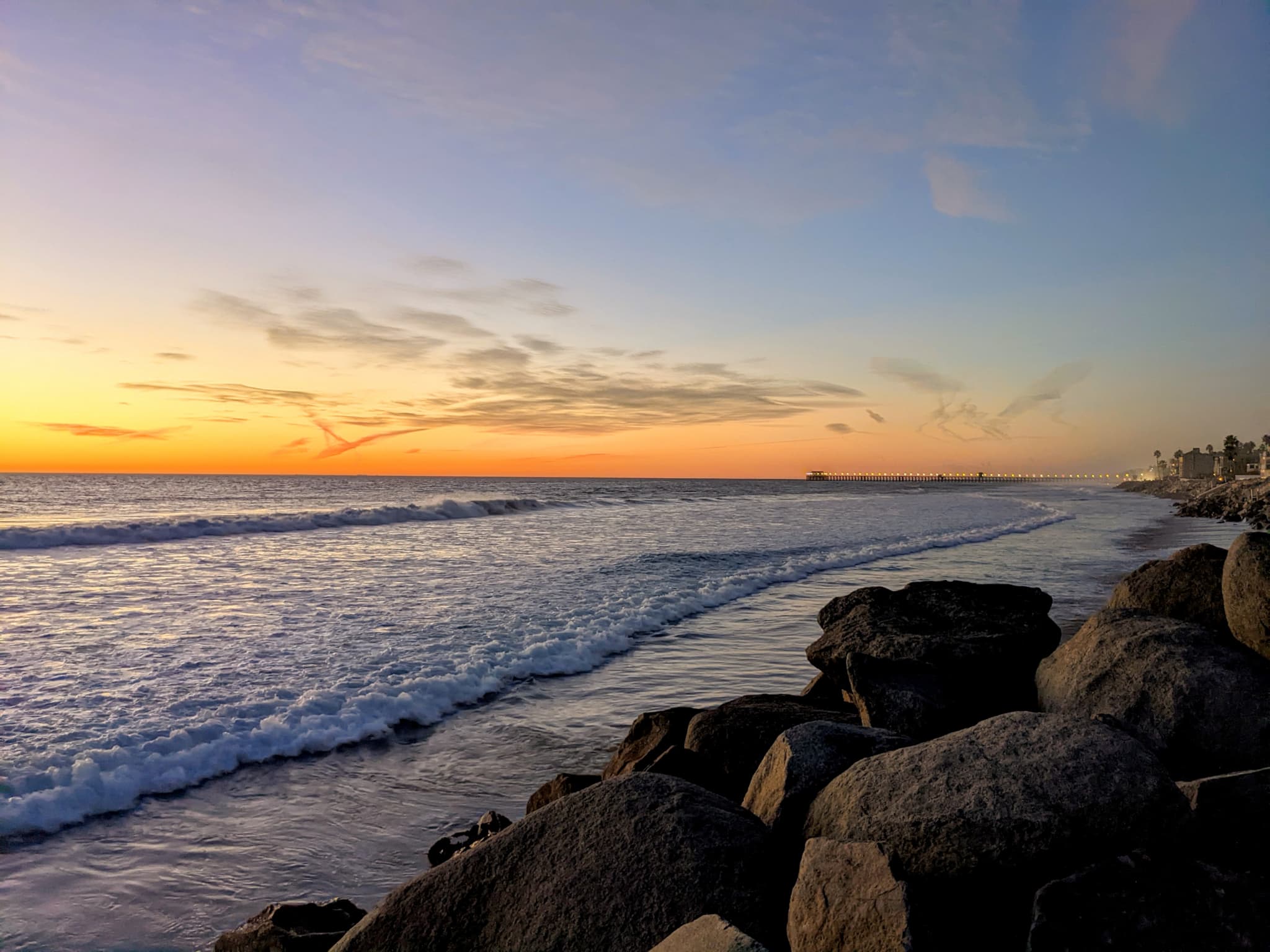 Orange sunset on a rocky shore with lit up pier in background.