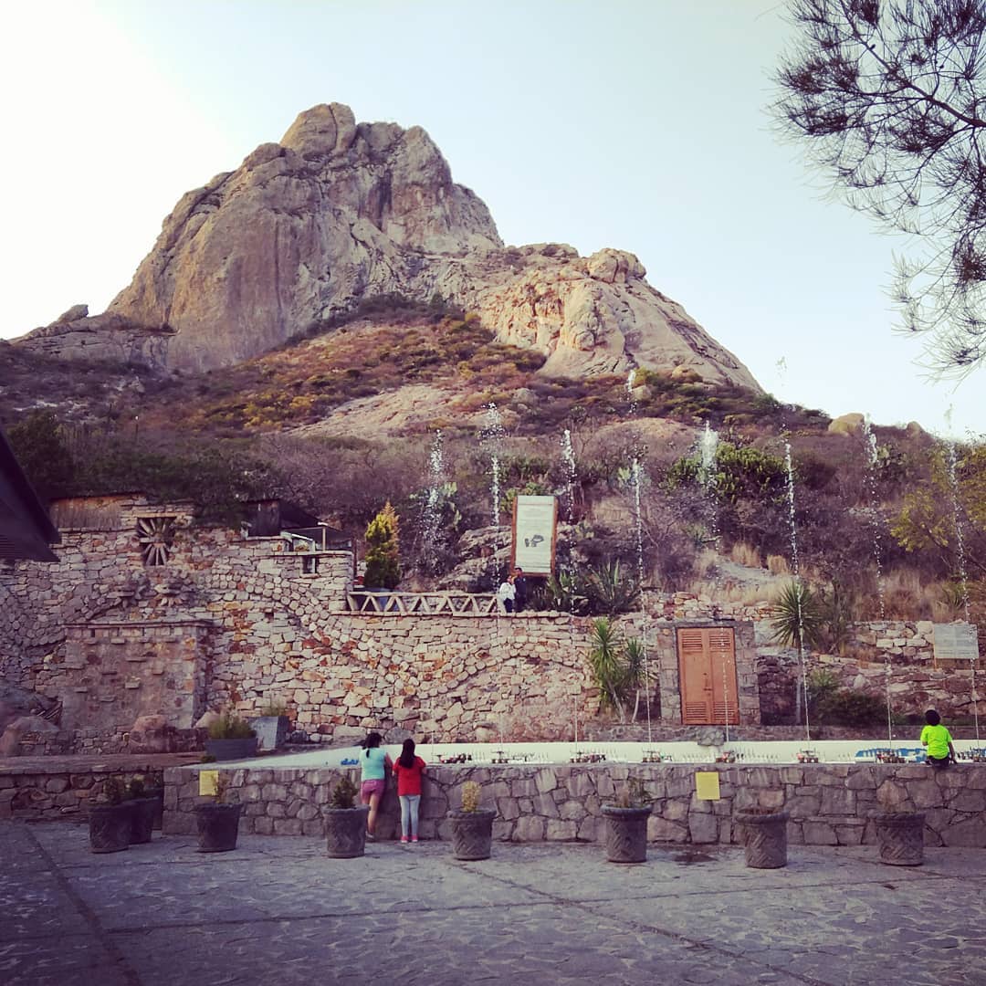 Looking up at a large rock formation. Below a large retaining wall, two children look at a water fountain.