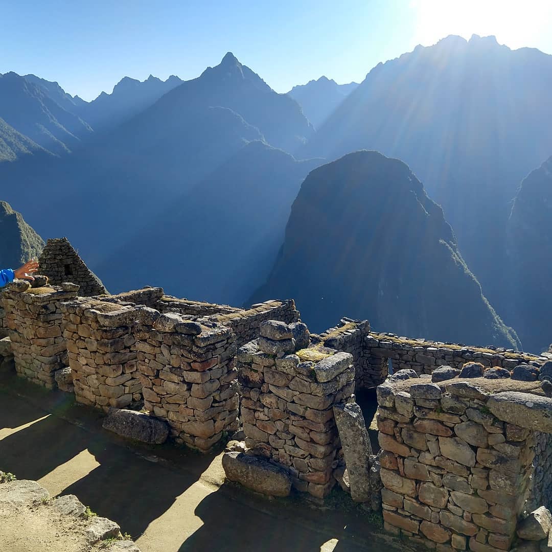 The sun shines over the jagged peak of mountains towards the stone walls of an abandoned settlement.