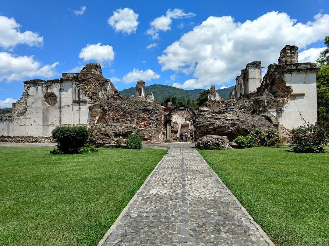 Concrete path through a green lawn, leading to the ruins of a large building. An archway leading to a staircase still stands with walls collapsed around it.