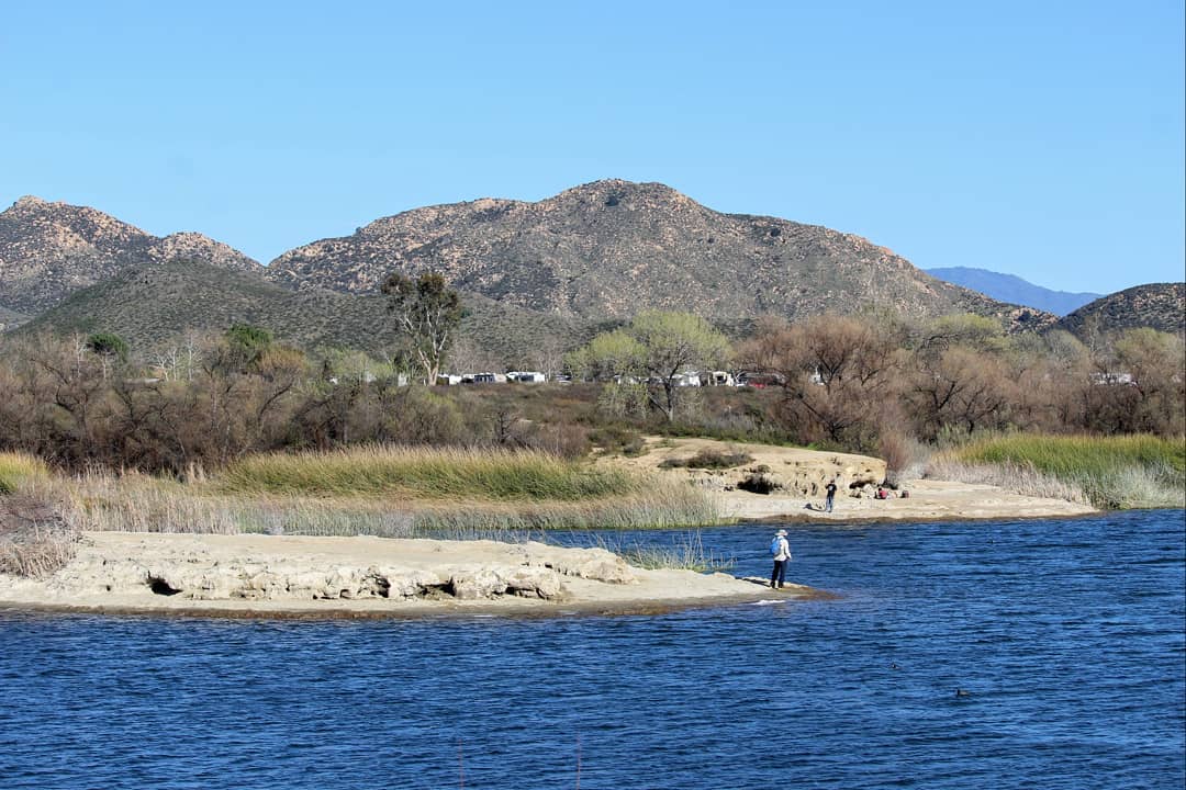 A scrubby, dry, shoreline, next to deep blue water with rocky hills behind.