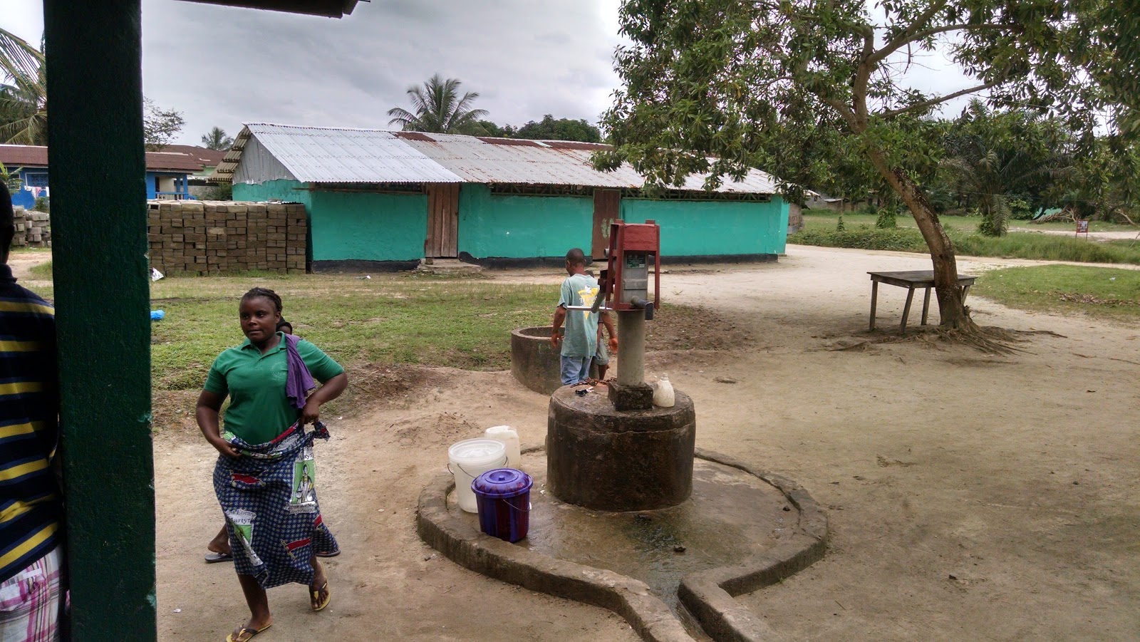 dirt yard with a tree and a manual water pump and a brightly colored building in the background