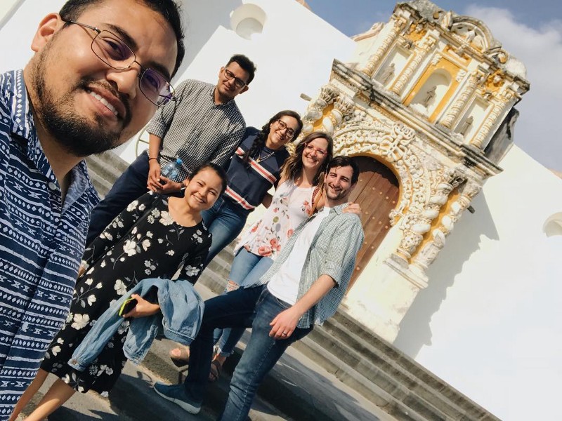 6 young people, 4 mexican, 2 white taking a selfie in front of a white building with a decorated entryway