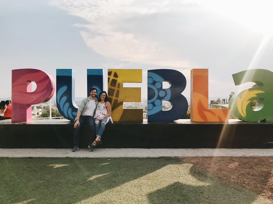 Colorful letters mounted to the ground spelling puebla with a couple sitting on the base